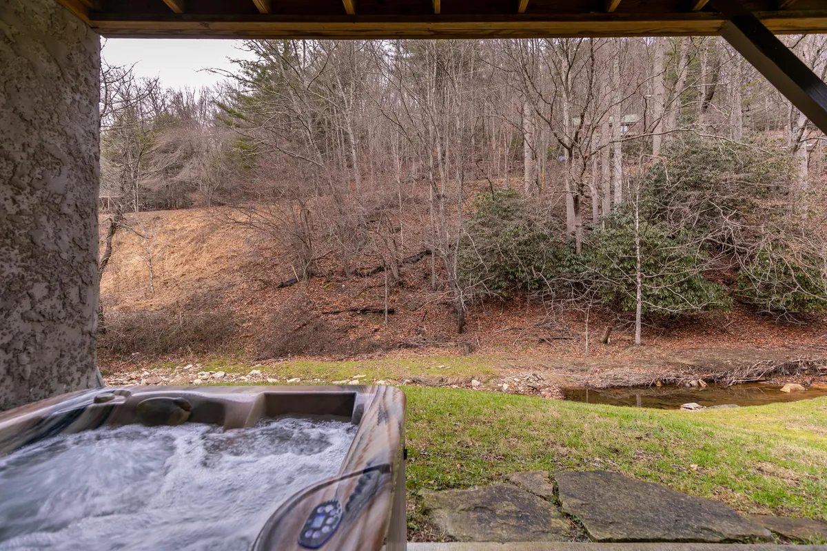 A serene outdoor patio setting with a bubbling hot tub overlooking a tranquil natural landscape and a flowing stream. The ambiance is peaceful, perfect for unwinding and enjoying nature.