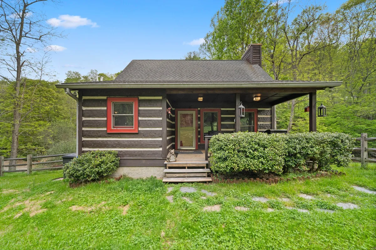 A welcoming log cabin with charming red window frames and a spacious front porch, set amidst landscaped greenery. The porch beckons for morning coffee or an evening glass of wine while surrounded by mountain nature.