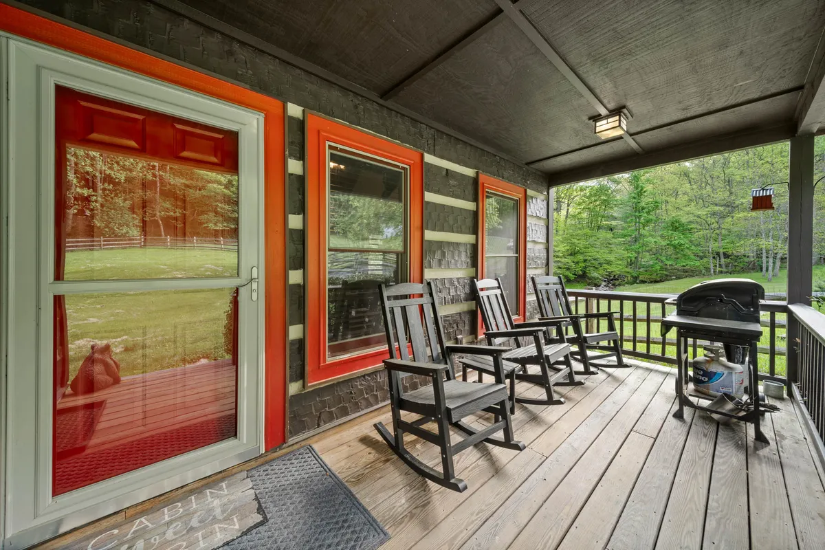 The front porch of the cabin features classic rocking chairs and a grill, framed by a vibrant red door. The porch setting is inviting and suggests opportunities for outdoor dining, grilling, and stargazing, set against the cozy backdrop of the cabin’s entrance.