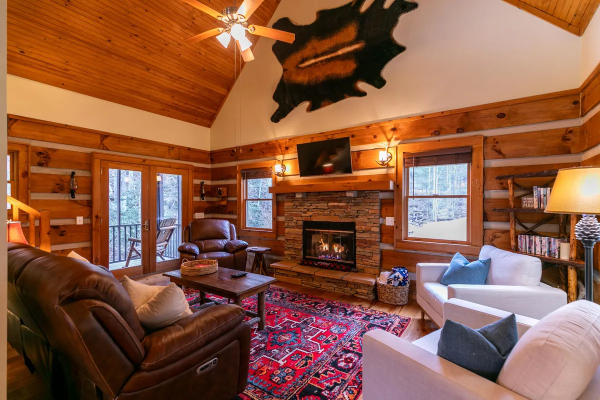 A spacious living area with a central stone fireplace, comfortable seating, exposed wooden beams, cozy rug, and expansive windows framing serene mountain and greenery views. The arrangement invites gatherings, conversation, and relaxation.