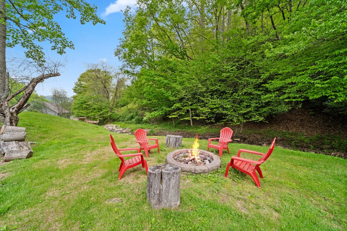 A cozy fire pit encircled by inviting red Adirondack chairs, nestled among lush green foliage. The setup is perfect for group gatherings, with space for storytelling and relaxation as evening falls. The surrounding natural beauty and communal seating make it an ideal spot for sharing memories under the stars.