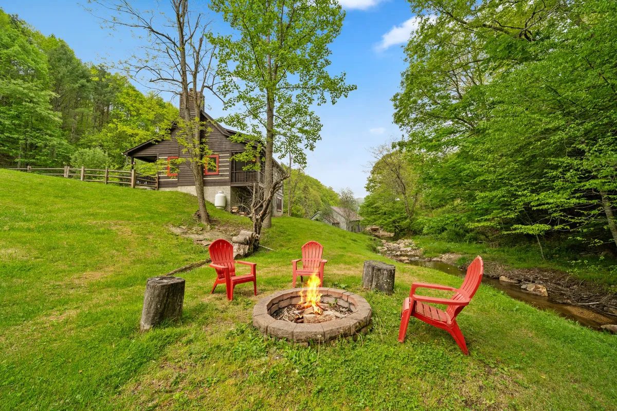 A cozy fire pit scene encircled by red chairs, set amidst greenery with the cabin visible in the background, perfect for evening campfires and storytelling under the stars.
