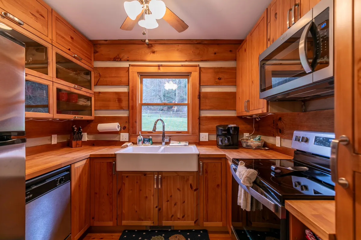 This image showcases a warmly lit kitchen with wooden cabinetry, a large farmhouse-style porcelain sink, and a window that lets in natural light. The kitchen appears inviting and functional, emphasizing its suitability for cooking and gathering.