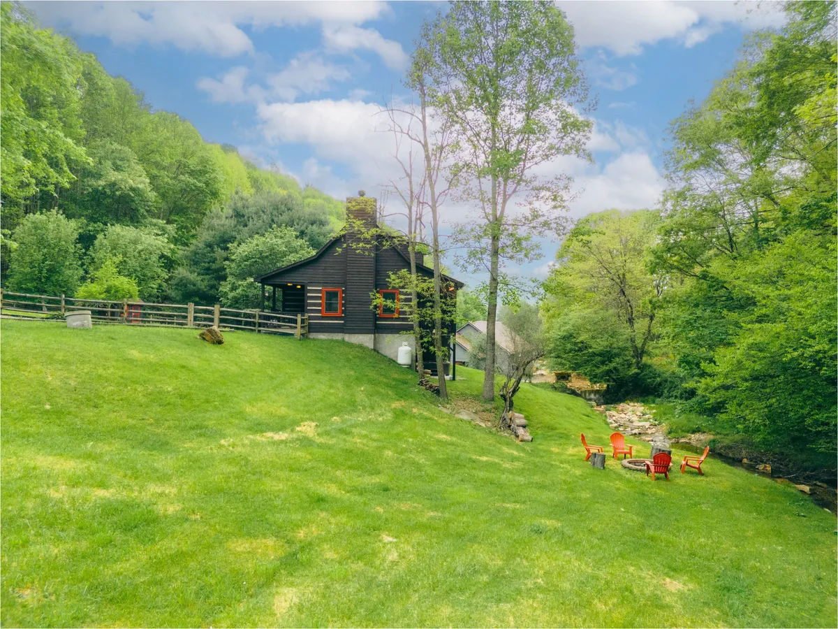The image shows the cabin beside a lively stream, with red chairs arranged around a fire pit. The green trees and calm water create a peaceful setting for relaxing, playing, or gathering in the evenings for s'mores and stories.
