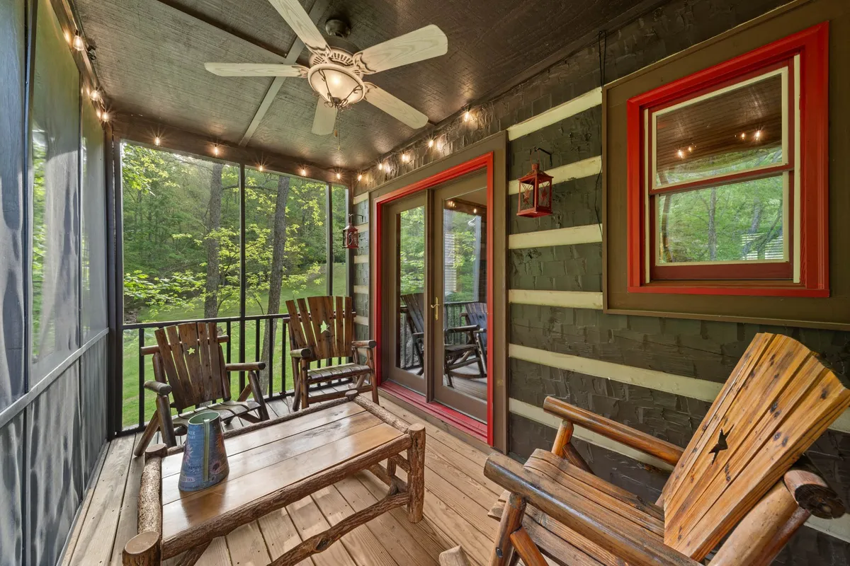 A cozy screened-in porch furnished with wooden rocking chairs, a rustic wooden table, a hanging lantern, and a ceiling fan. The space is framed by screens that offer views into the surrounding greenery, making it ideal for enjoying coffee or conversation while immersed in nature.