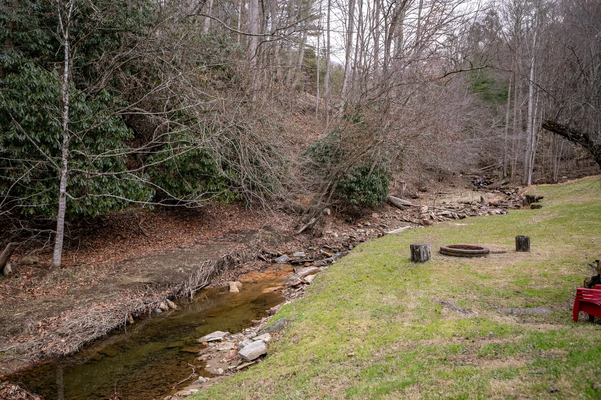 A peaceful outdoor scene featuring a clear stream running alongside a grassy area dotted with trees. A fire pit sits near the water, providing a cozy spot for gathering. The natural setting is perfect for relaxing, fishing, or exploring the surrounding landscape.