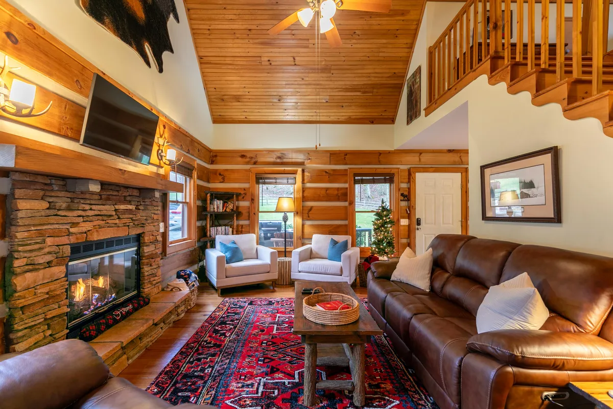 This image features a cozy living room with a rustic stone fireplace, a plush leather sofa, wooden ceiling beams, an inviting area rug, and armchairs. Large windows let in soft natural light, subtly hinting at the surrounding mountain views.