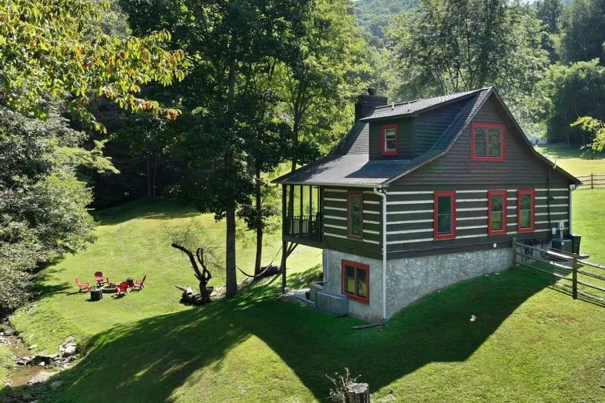 The image features a charming log cabin with red accents nestled along a peaceful creek. The outdoor seating area invites guests to unwind, while the golden glow of sunset bathes the screened porch in warm light. The natural surroundings evoke tranquility, with the promise of stargazing and bonfires under the clear Banner Elk sky.