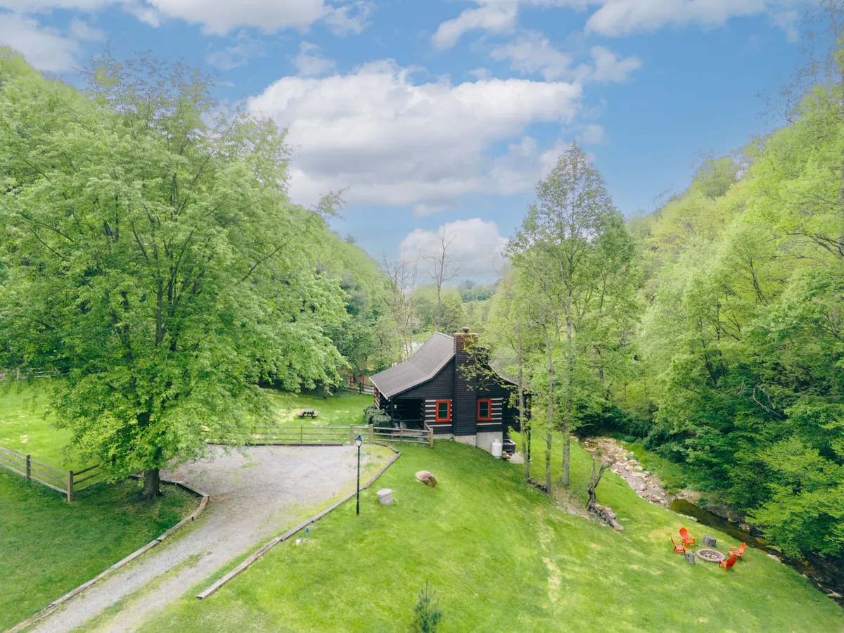 A scenic view of the log cabin tucked beneath large trees, with a gentle stream running alongside and red chairs placed nearby for riverside relaxation. The tranquil environment is perfect for unwinding, enjoying nature, or gathering with friends after a day of local adventures.