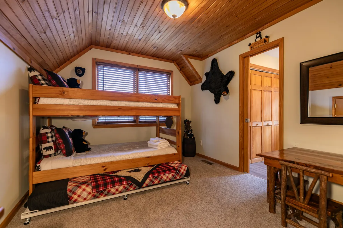 A loft space showcasing twin bunk beds with textured wood walls and ceiling, a small wooden table, and a playful bear decoration. The atmosphere is cheerful and inviting, ideal for children or family sleepovers.