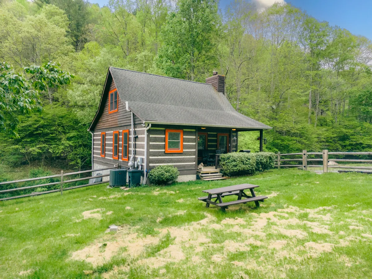 A welcoming view of the log cabin exterior, surrounded by lush greenery and featuring a brightly painted window trim. A picnic table sits in the foreground, inviting guests to dine or relax outdoors. The yard is spacious, providing ample room to enjoy fresh mountain air.