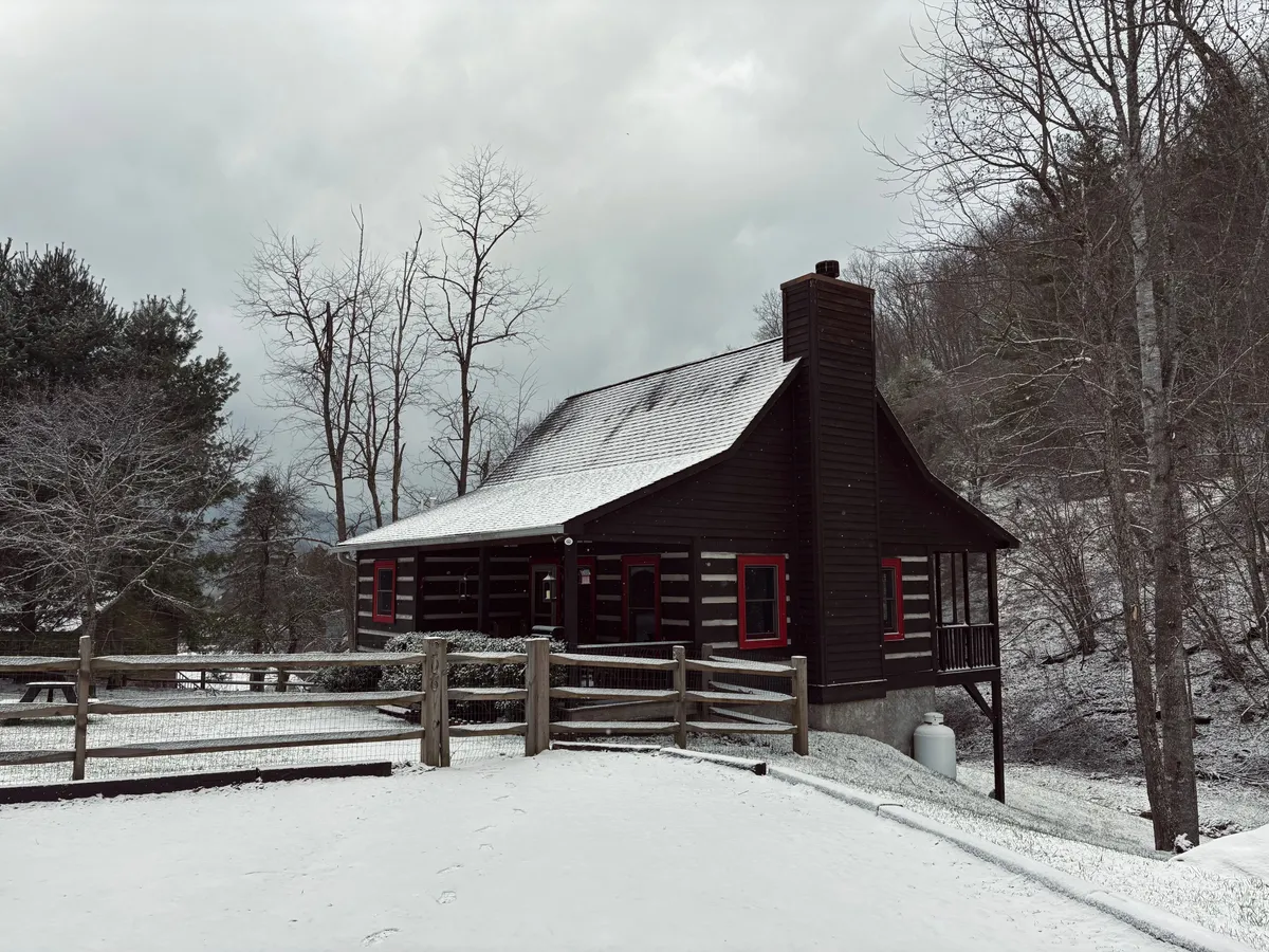 A tranquil winter scene with the log cabin nestled among snow-covered ground and surrounded by tall trees. The red window frames of the cabin pop against the white snow, evoking a cozy, secluded mountain getaway ideal for snowy adventures.