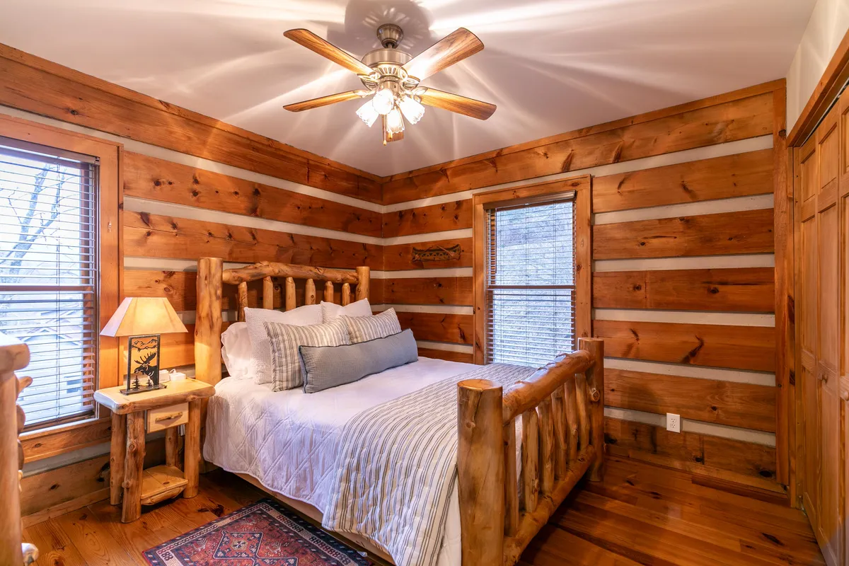 A cozy queen bedroom featuring a log bed with a plush comforter, a nightstand with a glowing lamp, a window letting in soft natural light, rustic wooden walls, and a warm area rug. The setting is intimate and designed for rest and tranquility.